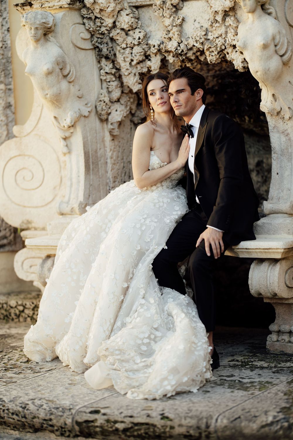 Romantic wedding couple sits together against ornate stone wall with elegant baroque architectural details.