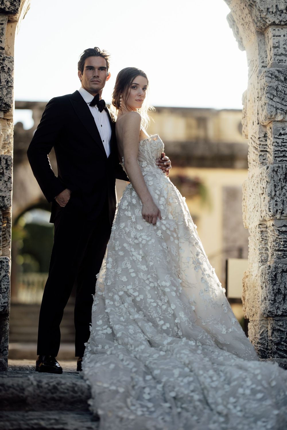 Elegant couple poses between stone pillars at sunset with bride wearing a flowing dress adorned with delicate floral details.