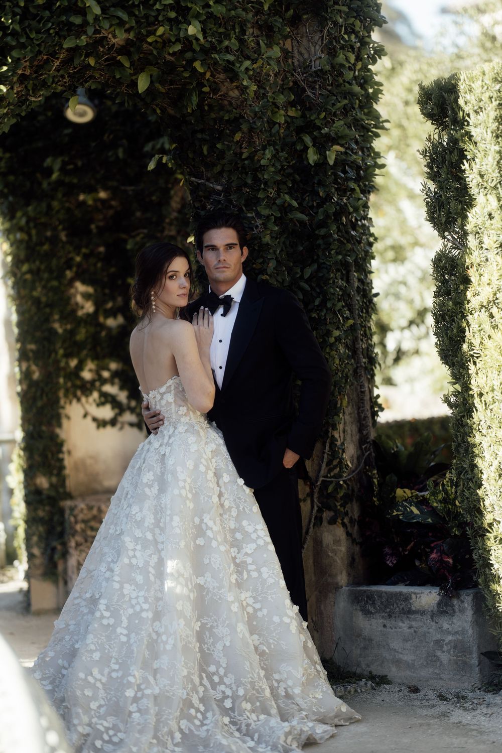 A bride in a floral applique ballgown poses with groom among garden greenery in an elegant outdoor wedding setting.