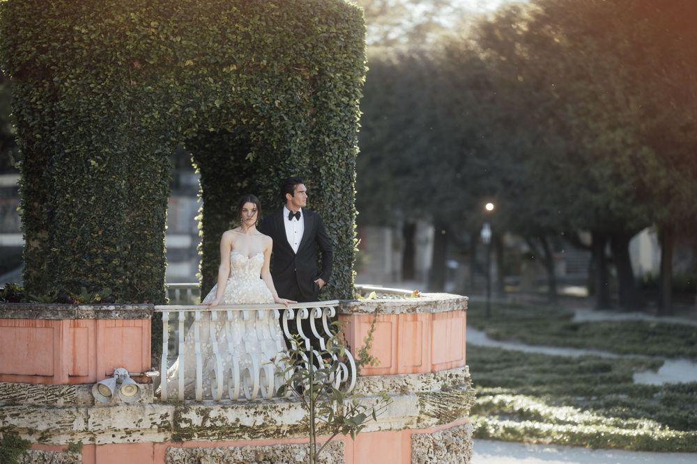 A romantic wedding couple stands at an ornate brick and wrought iron balustrade surrounded by manicured hedges at sunset.