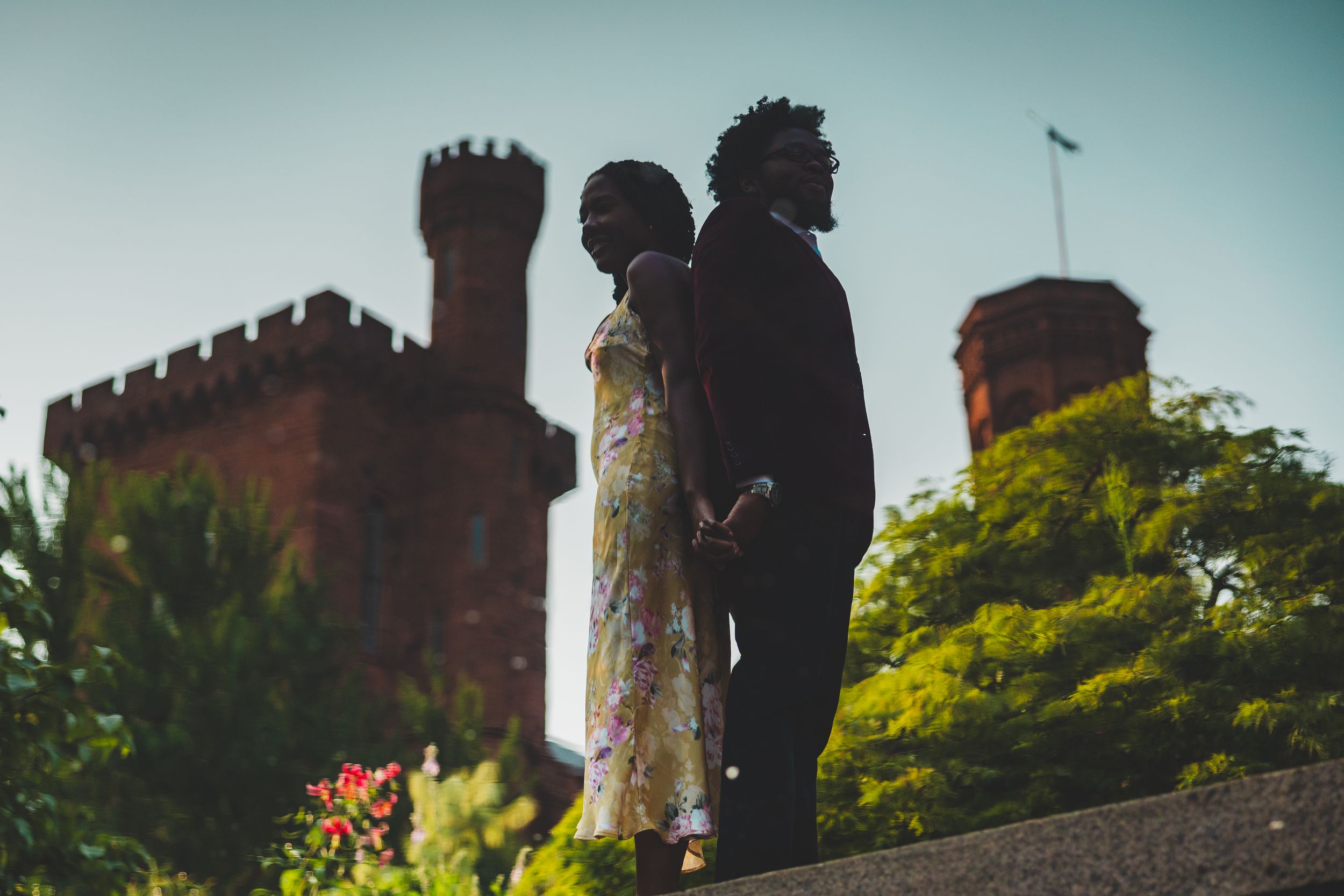 Silhouettes of two people standing together against a castle-like structure with turrets at sunset.
