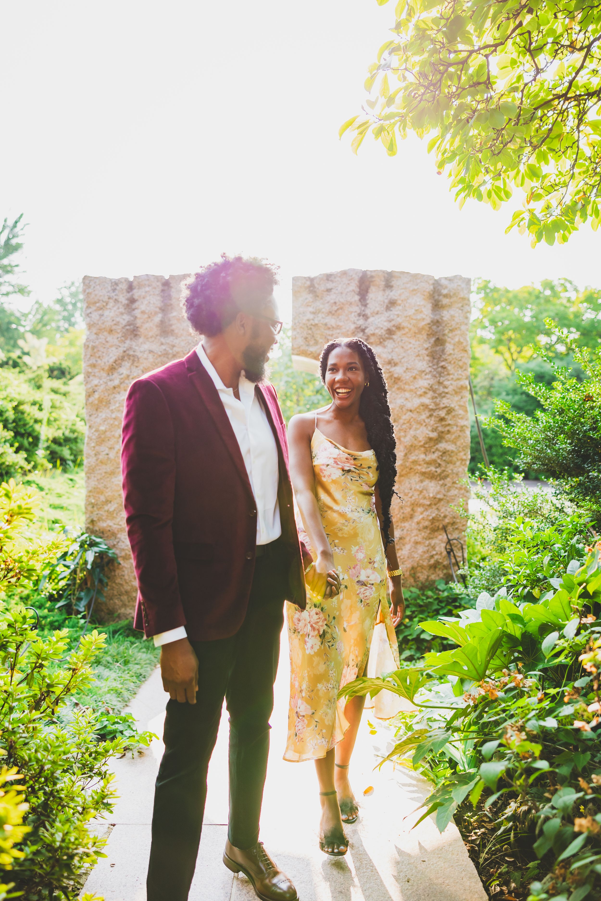 A couple walks together through a lush garden path with stone columns and dappled sunlight.