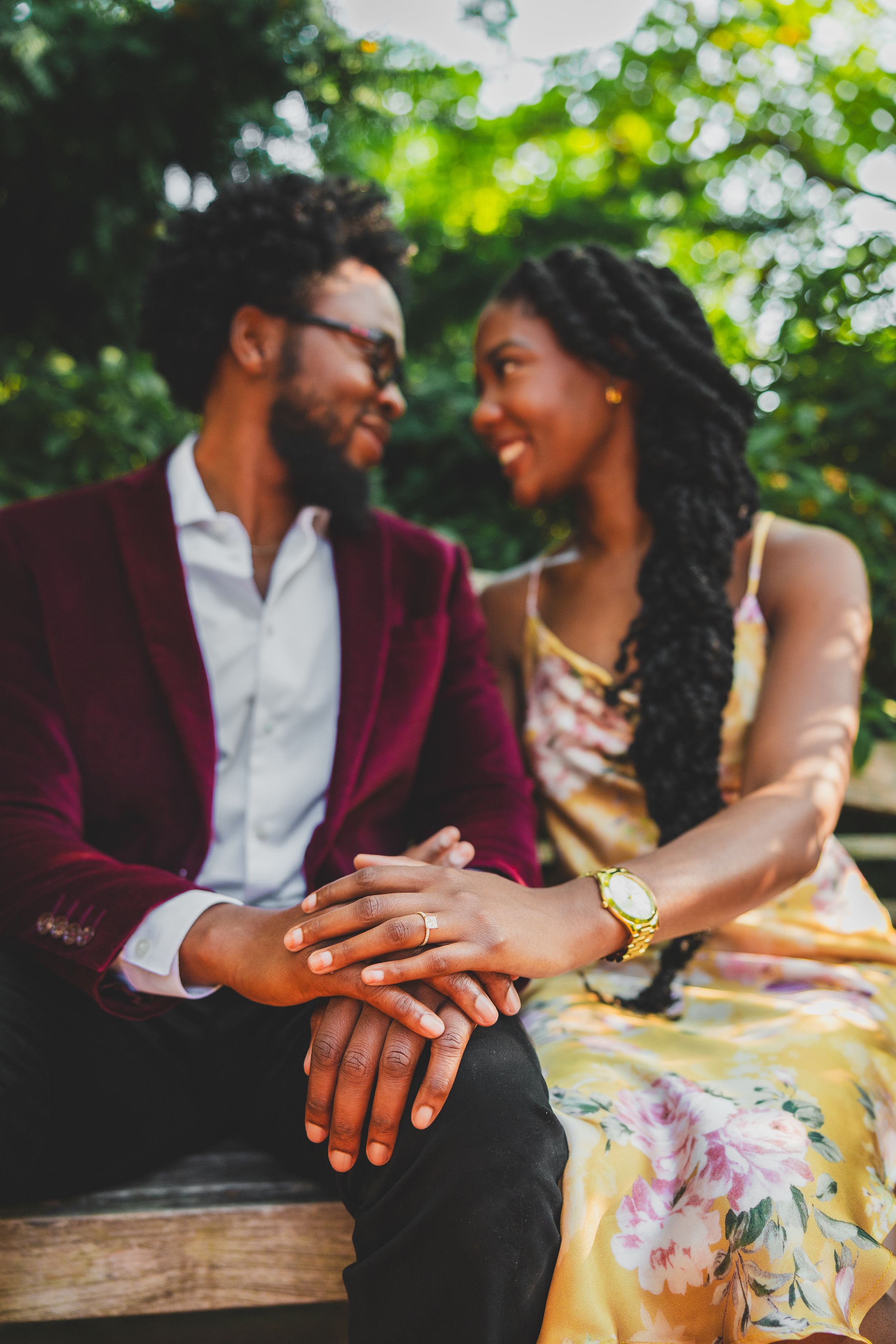 A romantic couple sits on a wooden bench in a garden setting, wearing burgundy and floral attire.