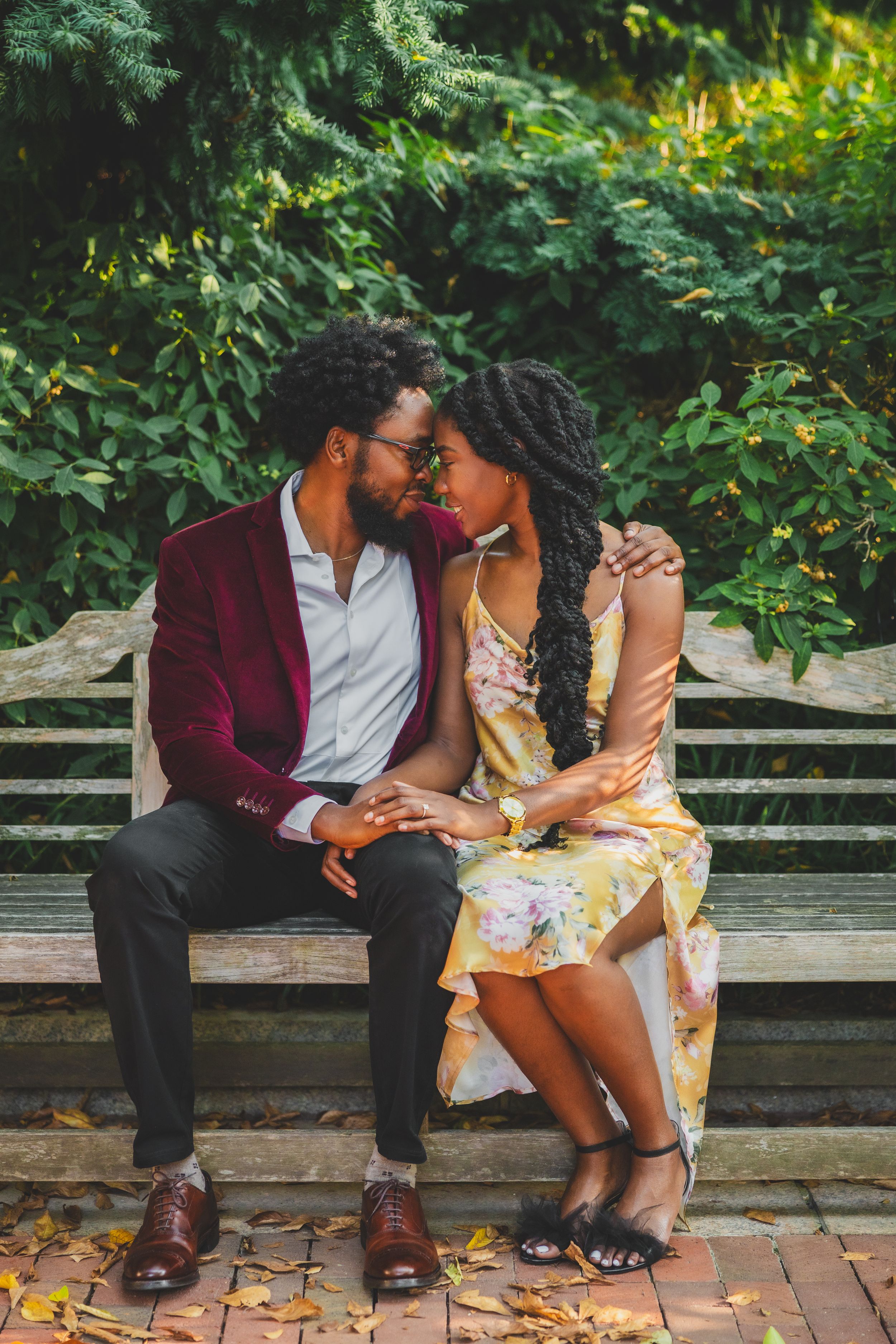 A romantic couple sits close together on a wooden bench surrounded by lush greenery and fallen autumn leaves.