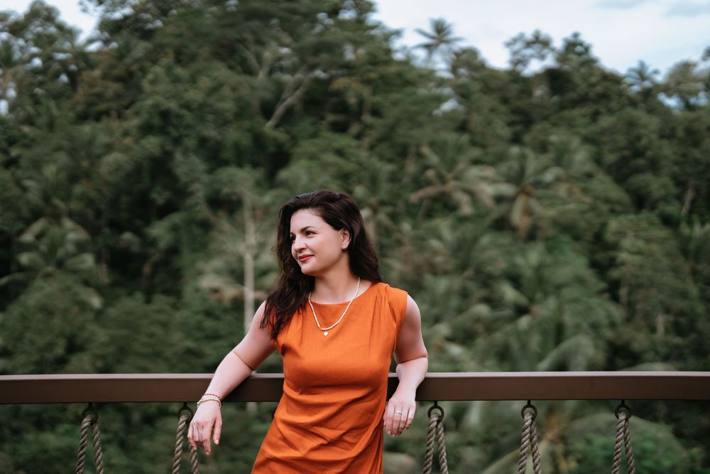 A figure in a bright orange dress poses against a lush forest backdrop with tall trees.