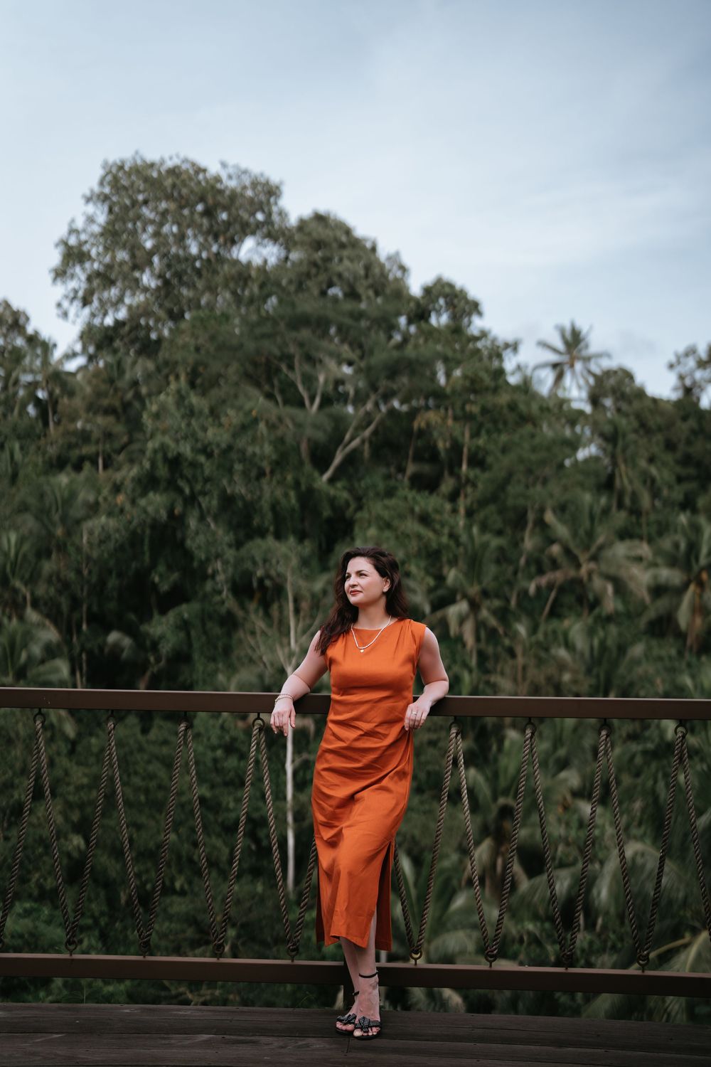 A person in an orange dress sits casually on a wooden railing with palm trees in the background.