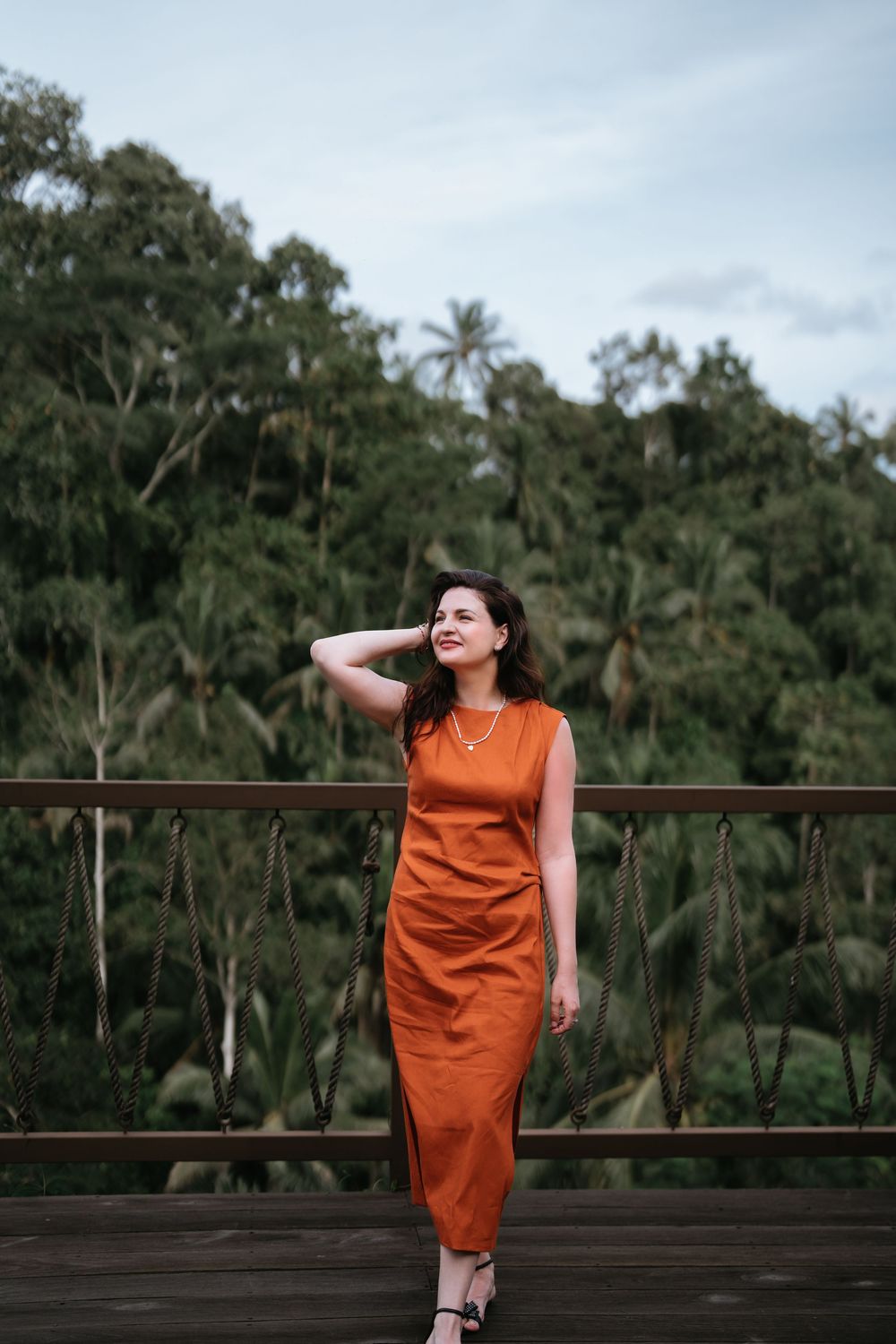 An orange-clad figure poses expressively on a wooden deck with tropical foliage in the distance.