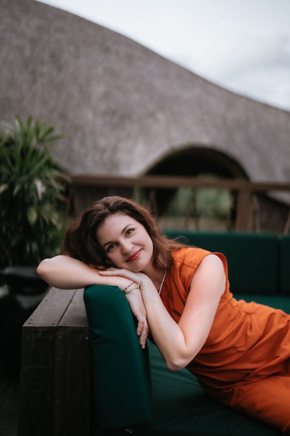 Relaxed lounging pose on dark furniture with tropical architecture visible.