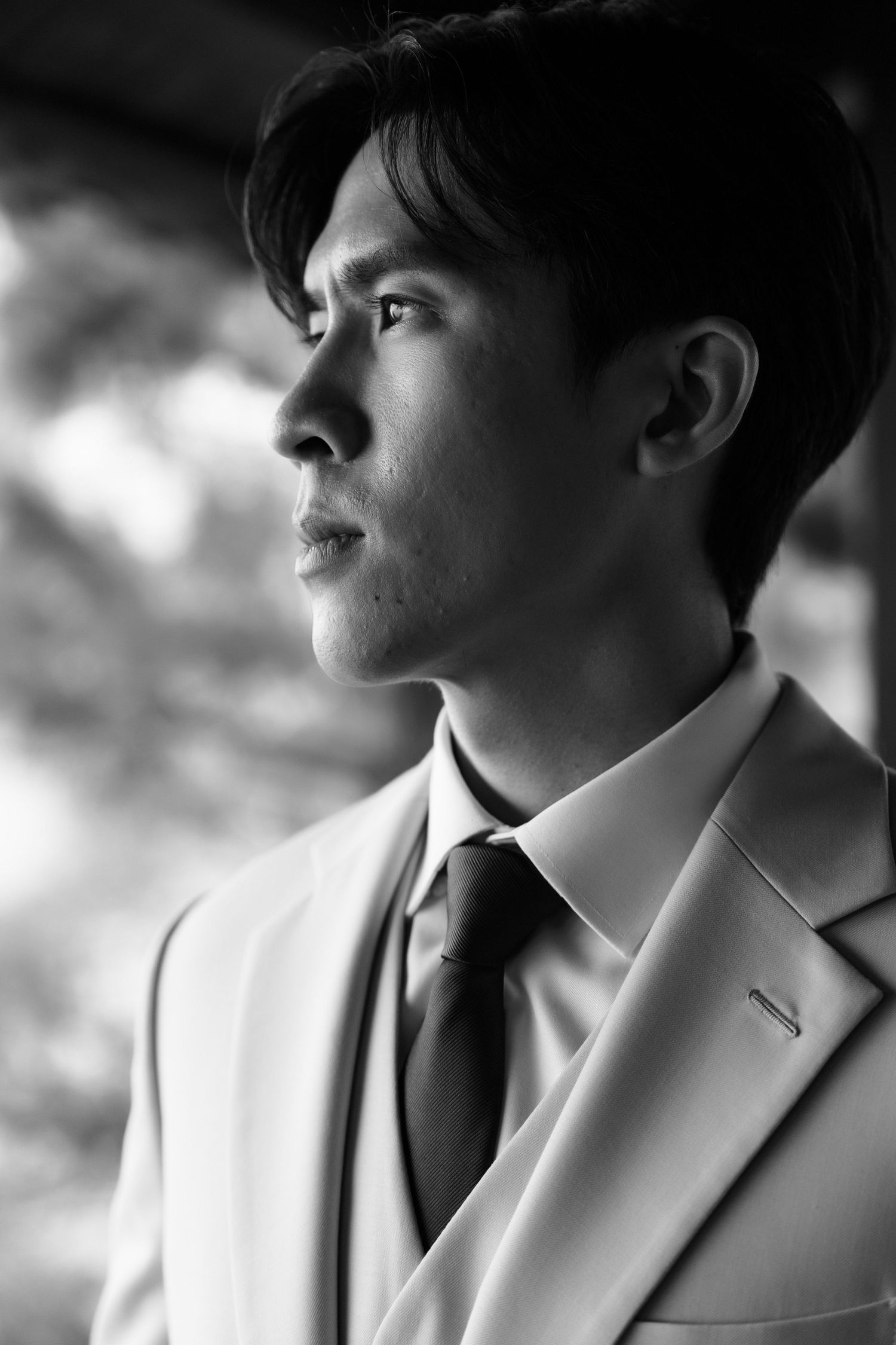 Black and white portrait of a young Asian man in a white suit and dark tie gazing thoughtfully to the side.