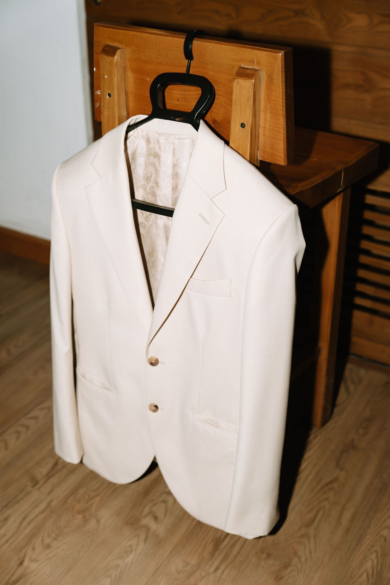 Elegant white men's blazer with gold buttons hanging on a black hanger against wooden furniture.
