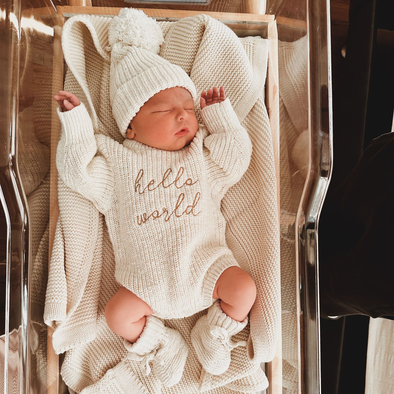 A newborn baby wearing a cozy cream-colored knit onesie and matching hat lies peacefully in a bassinet.
