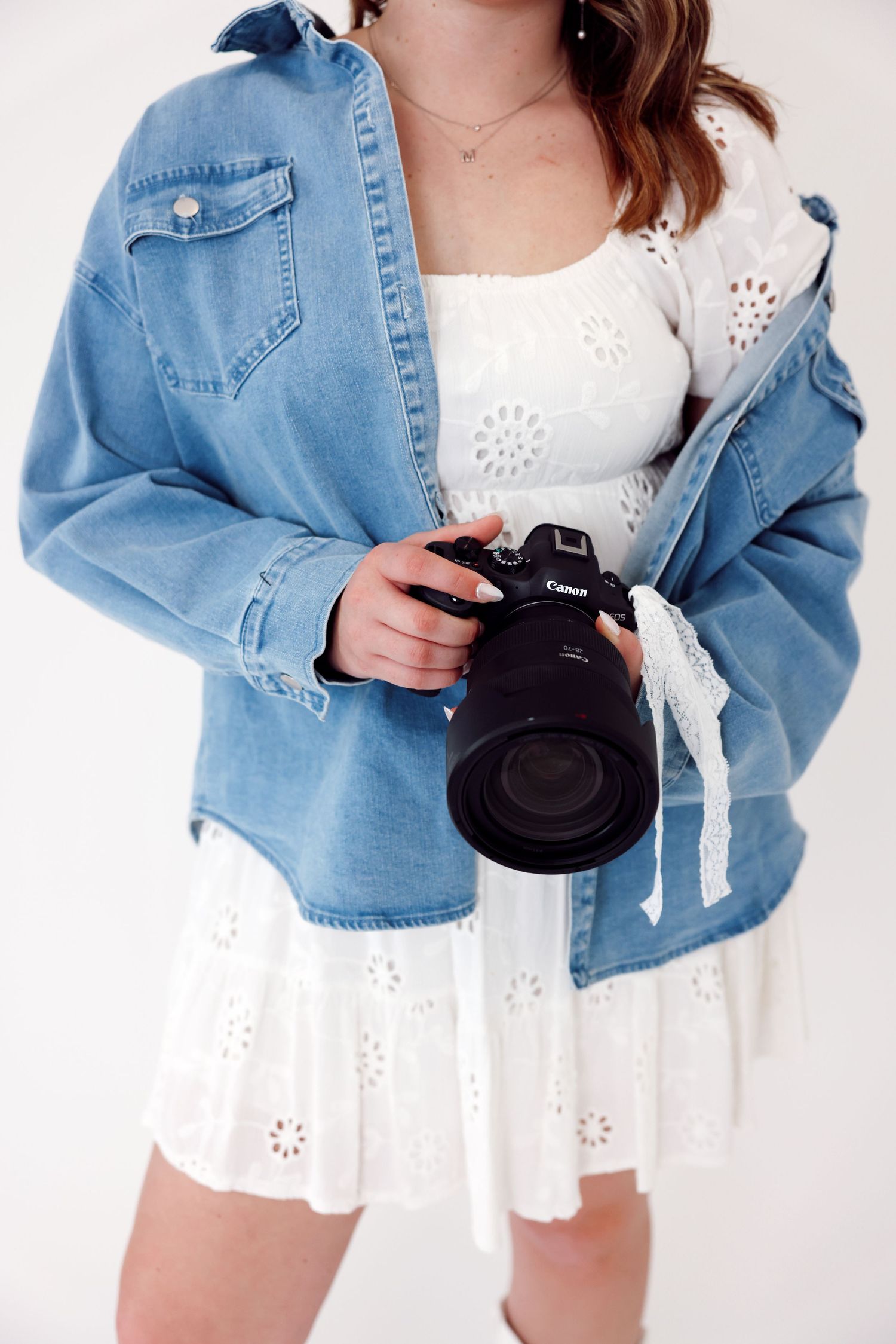 A photographer in blue overalls and white sweater holds a DSLR camera on their head while smiling against white background.