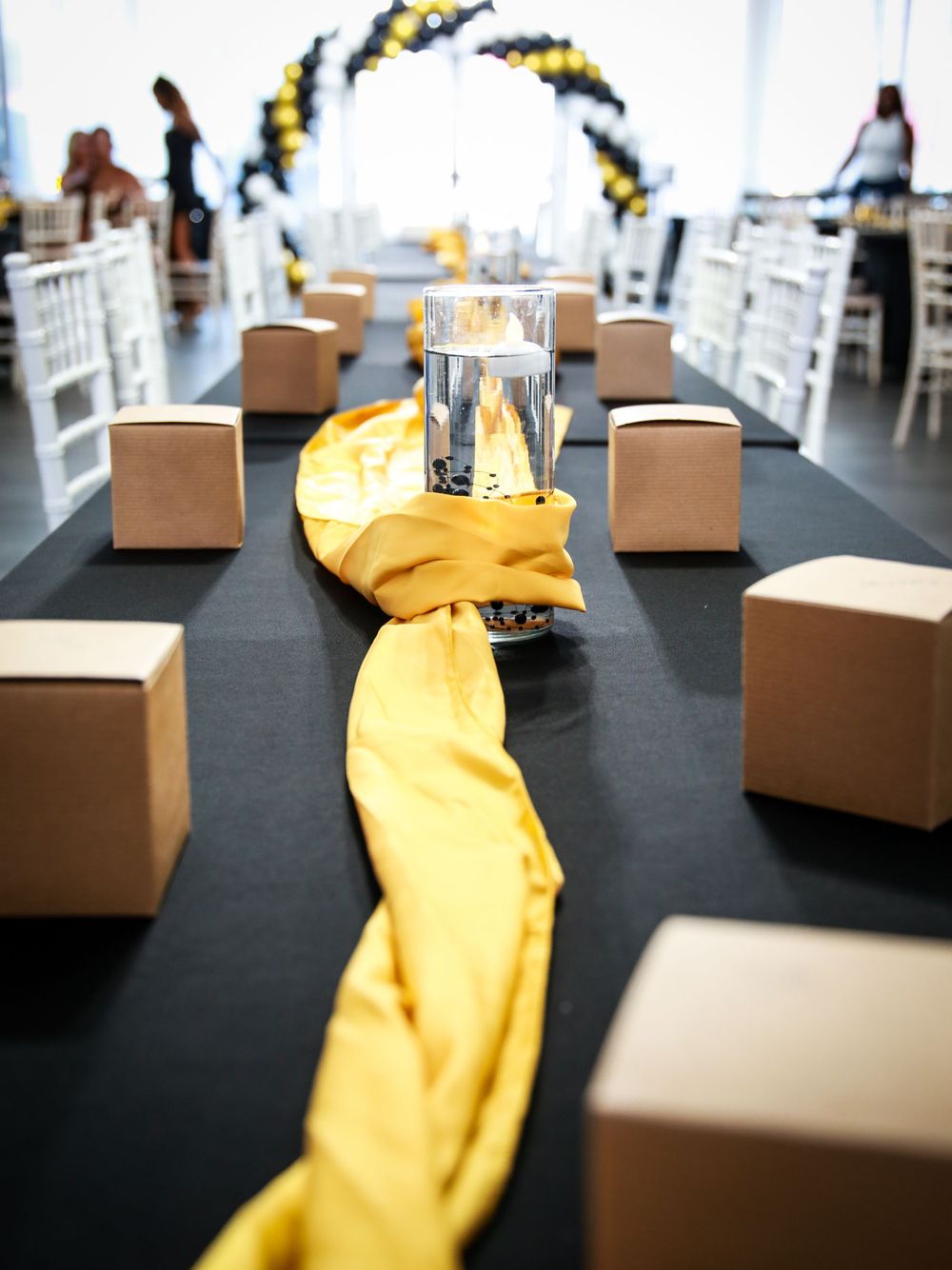Wedding reception table setup with black tablecloth, yellow runner, boxes for centerpieces, and balloon arch in background.