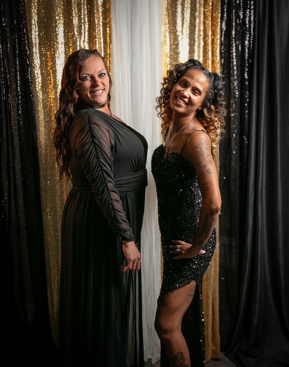 Two people posing together against a backdrop with gold and black sequin curtains in evening attire.