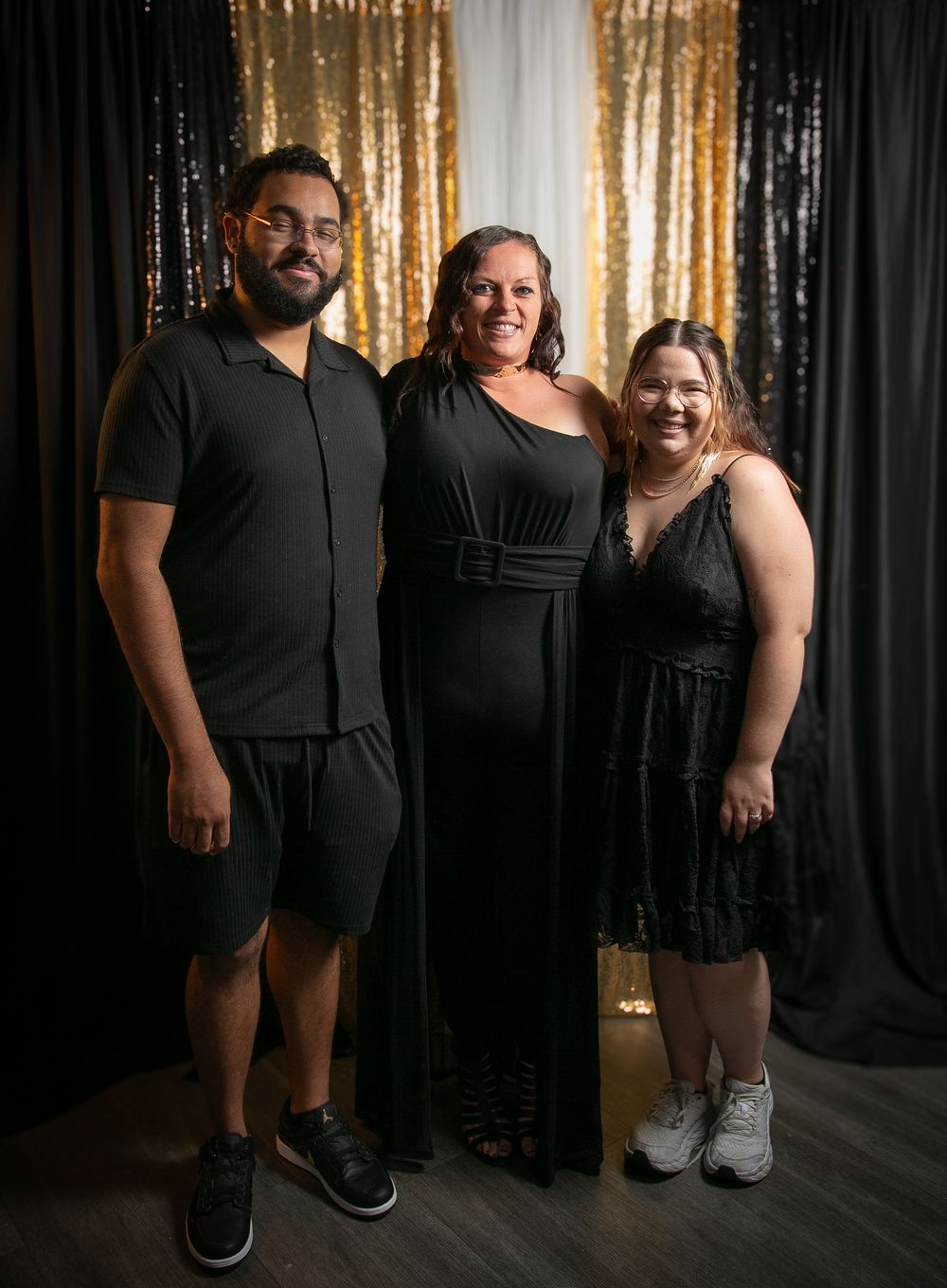 Three people wearing black outfits pose together against a backdrop with gold and black curtains.