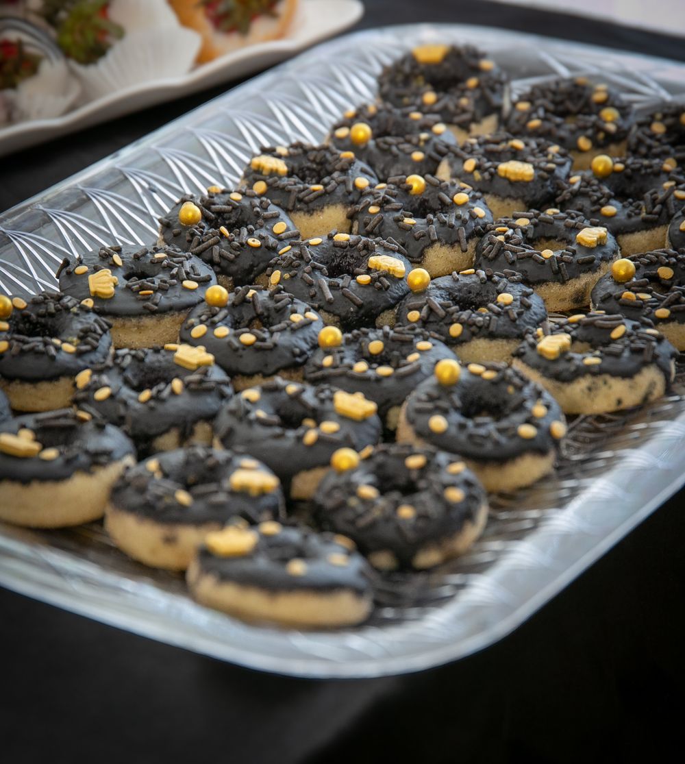 Gourmet chocolate donuts decorated with dark frosting, metallic sprinkles and gold accents on a silver serving tray.