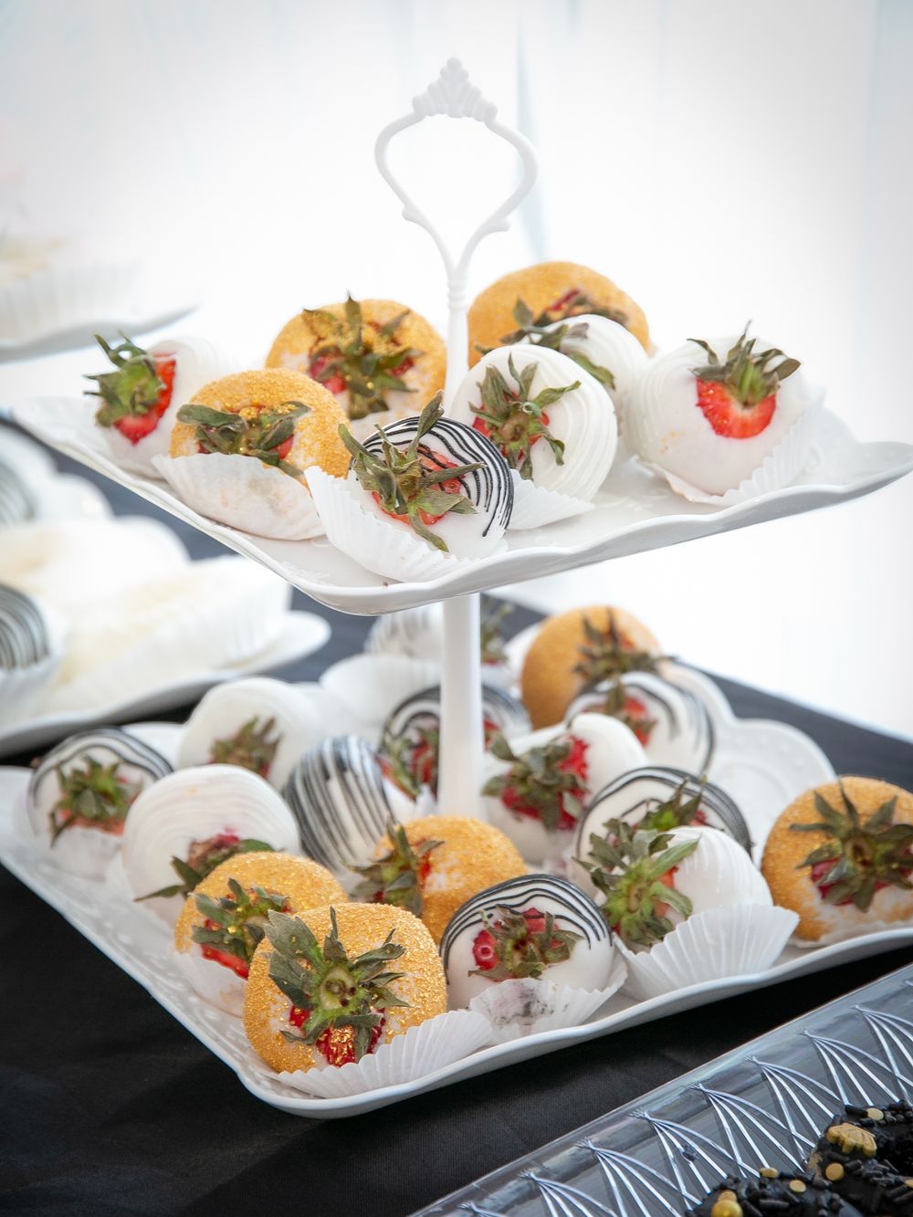 Chocolate covered strawberries and fruit displayed on a white tiered serving platter at an elegant dessert table.