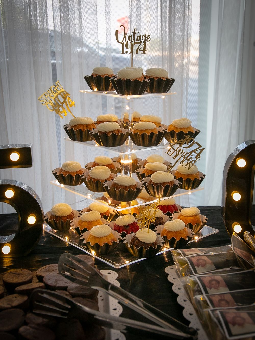 Multi-tiered display of frosted cupcakes with vintage inspired gold decorative toppers at a party setting.