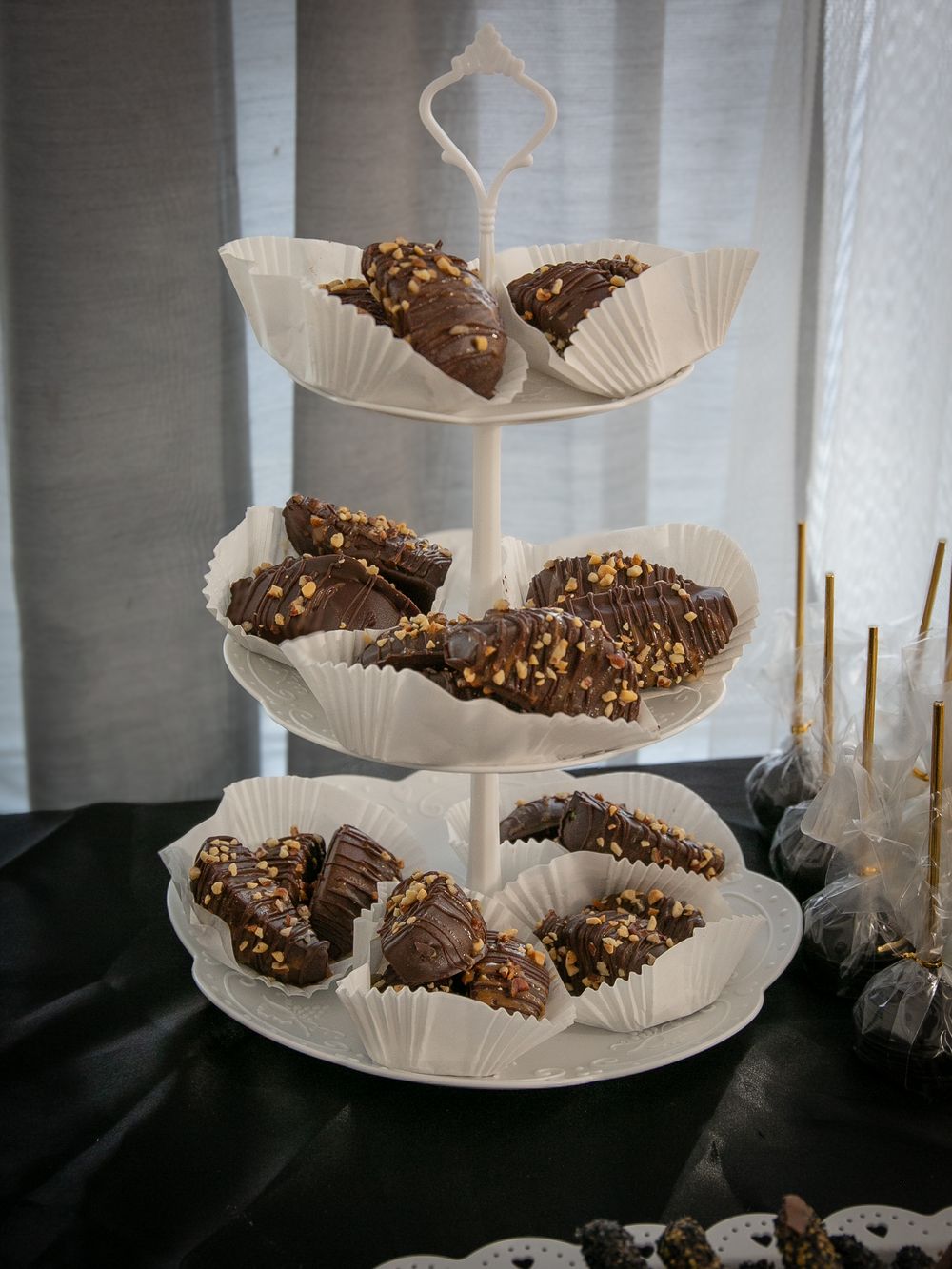 Three-tiered white serving stand displaying chocolate covered treats garnished with nuts on white paper liners.