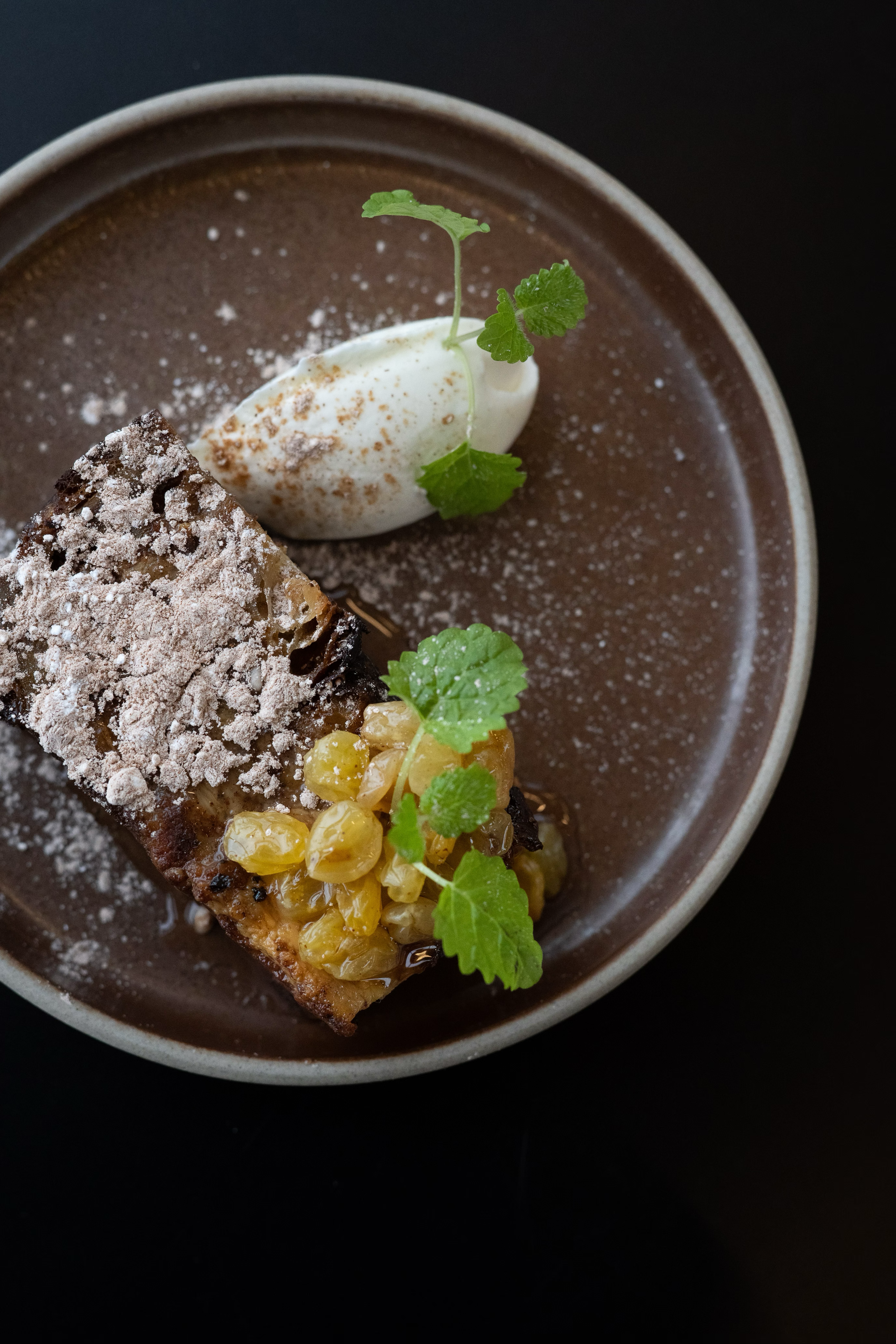 Dessert plate featuring chocolate crumble, whipped cream and microgreens on dark ceramic.