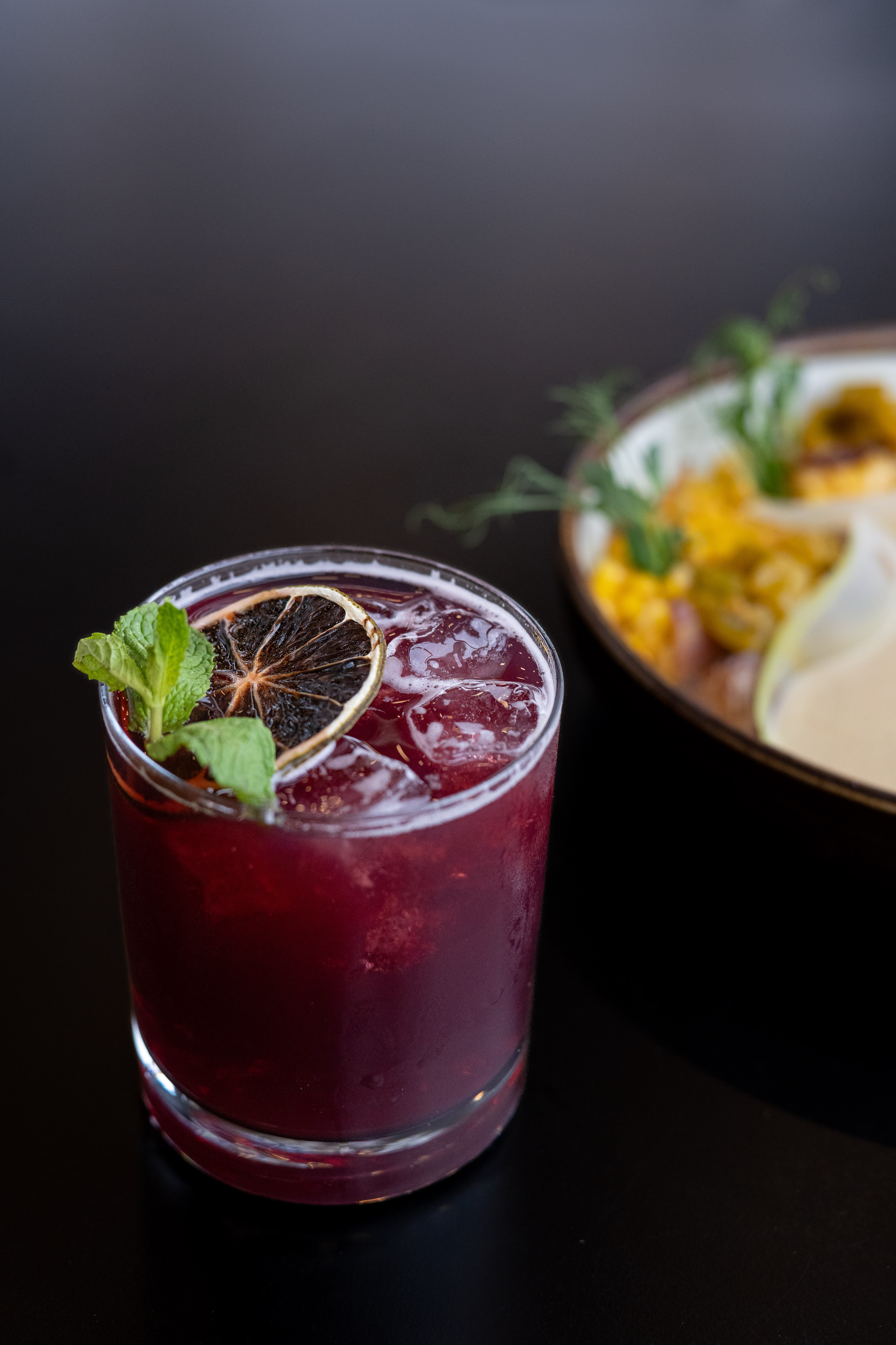 Deep red cocktail with ice, garnished with dried citrus and fresh herbs.