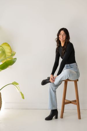 Lifestyle portraits of a person in casual attire sitting on a wooden stool against white backdrop with plant decor.