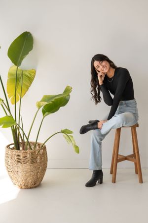 Studio photoshoot featuring casual poses on a wooden stool with decorative potted plant against white background.