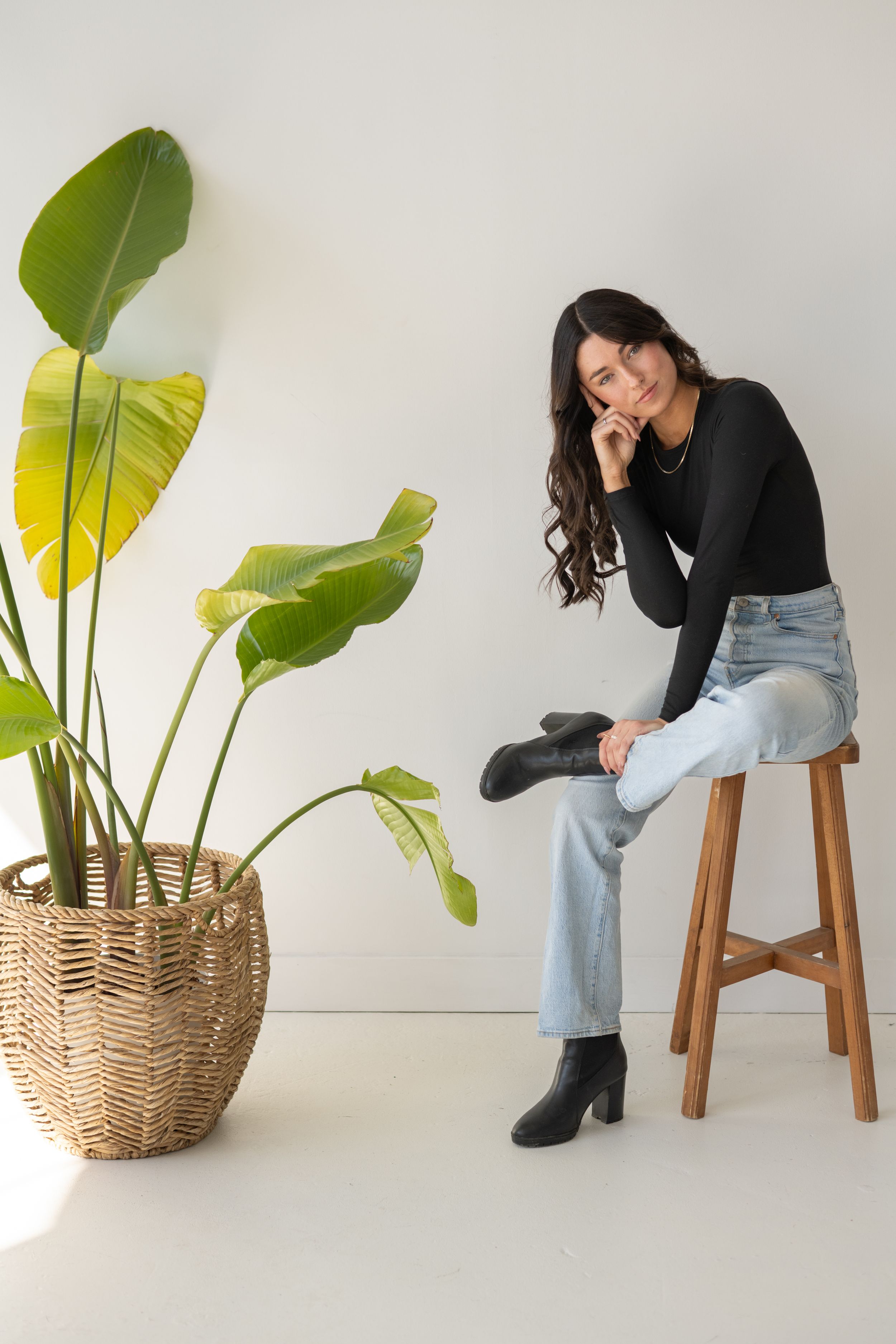 Creative portrait series with subject seated on wooden stool next to woven basket with large tropical plant leaves.