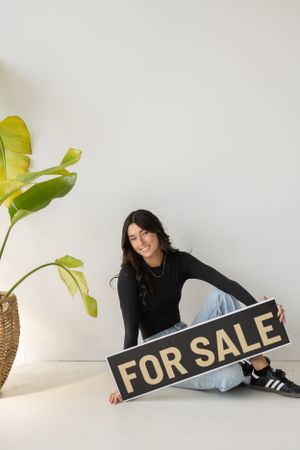 Lifestyle product photography scene with decorative plants and 'For Sale' sign against white wall.