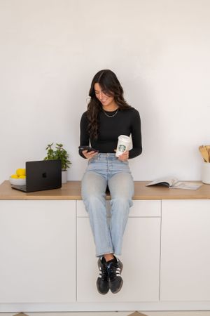 Casual workspace scene with someone in denim sitting on white cabinets with laptop and plants.