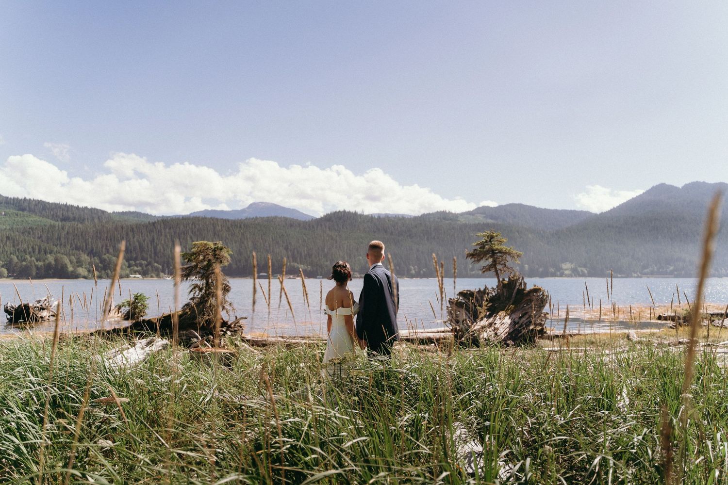 A distant couple stands by a mountain lake surrounded by scenic landscape and tall grasses.