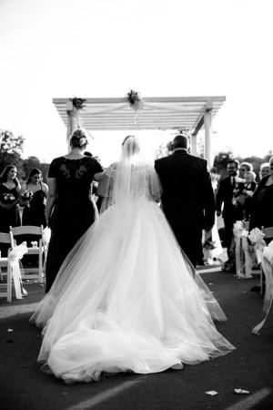 A bride in flowing white dress walks down the aisle under a white pergola during an outdoor ceremony.