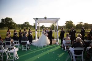 Outdoor wedding ceremony under white pergola with guests seated on white chairs.