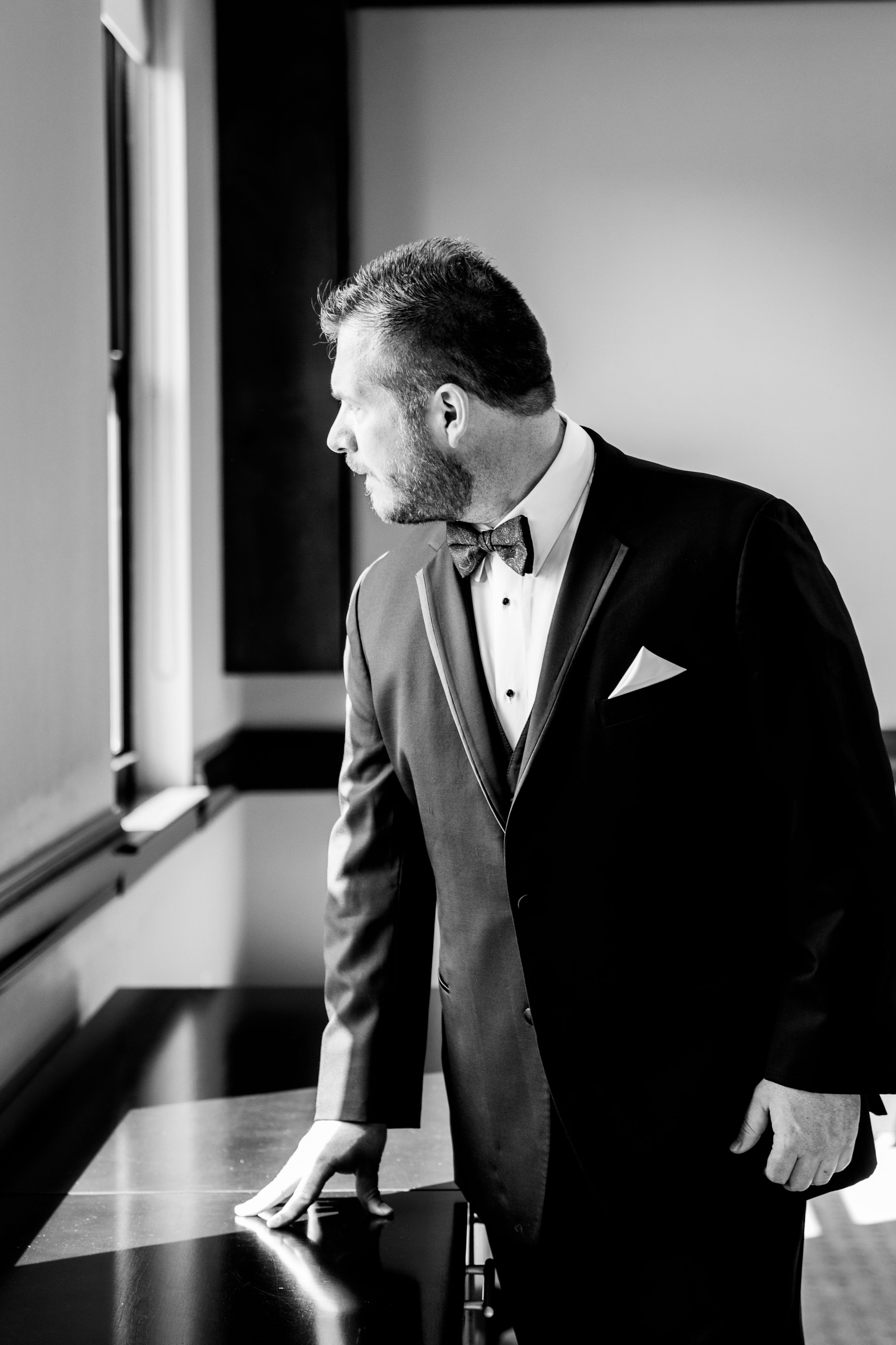 Side profile of a groom in formal tuxedo looking thoughtfully out a window.