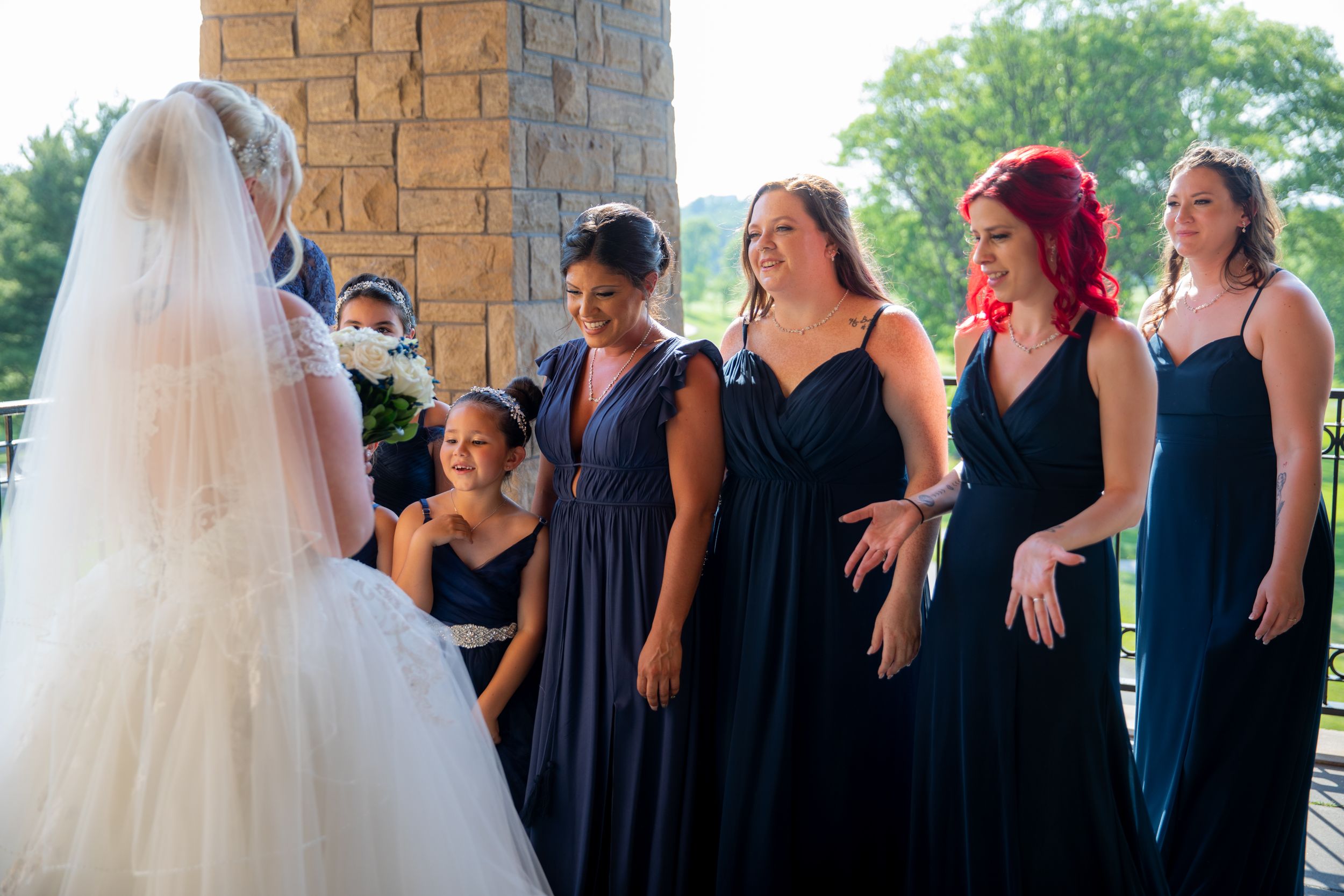 Bridesmaids in navy dresses react with joy seeing bride in her wedding gown.