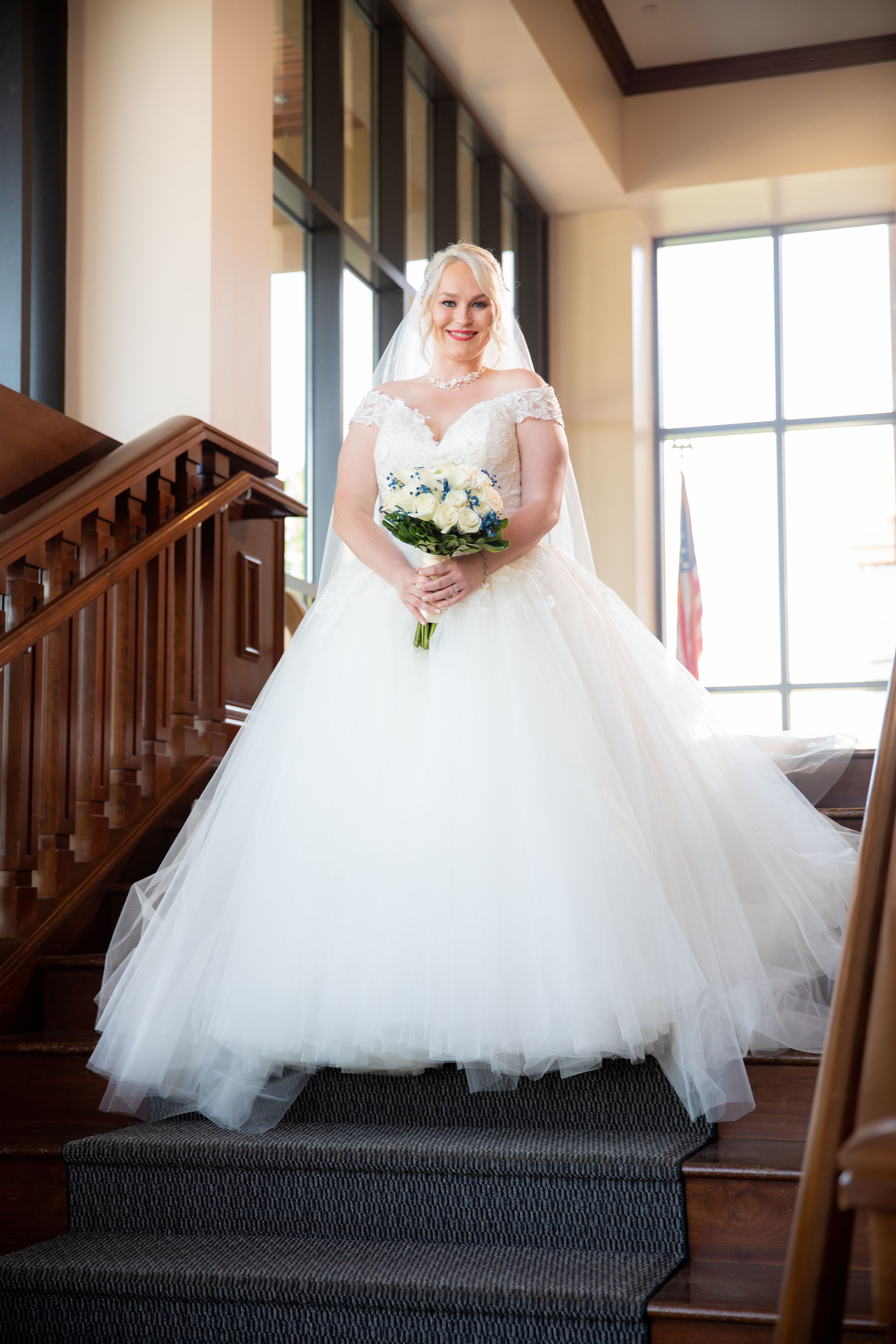 A bride in a stunning white ball gown with tulle skirt stands on carpeted stairs near a window.