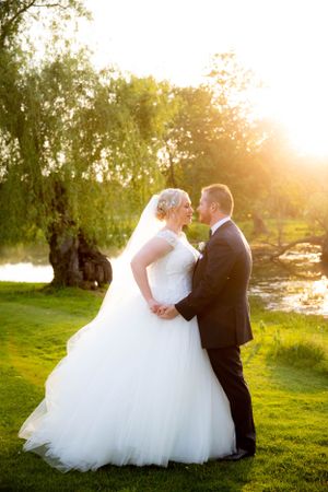 Romantic sunset photo of newlyweds standing on lush grass by a lake in warm golden light.