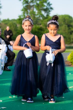 Two young flower girls in matching navy blue gowns carry baskets down an outdoor aisle.