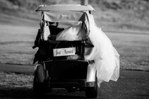 A wedding golf cart decorated with 'Just Married' sign carries a bride's flowing white dress down a path.