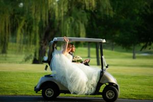 A bride in a flowing white dress sitting in a golf cart on a lush green course.