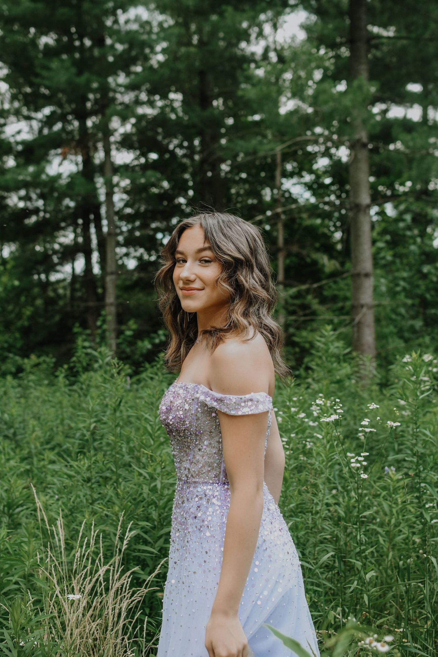 A dreamy off-shoulder lavender gown sparkles in a lush forest setting with soft natural lighting.