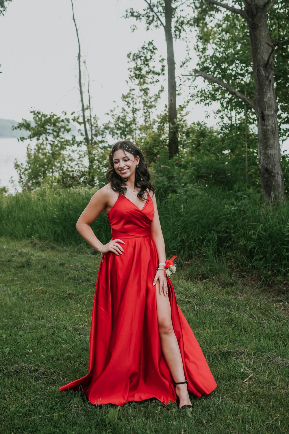 Elegant red formal gown with side slit photographed outdoors with trees and water in background.