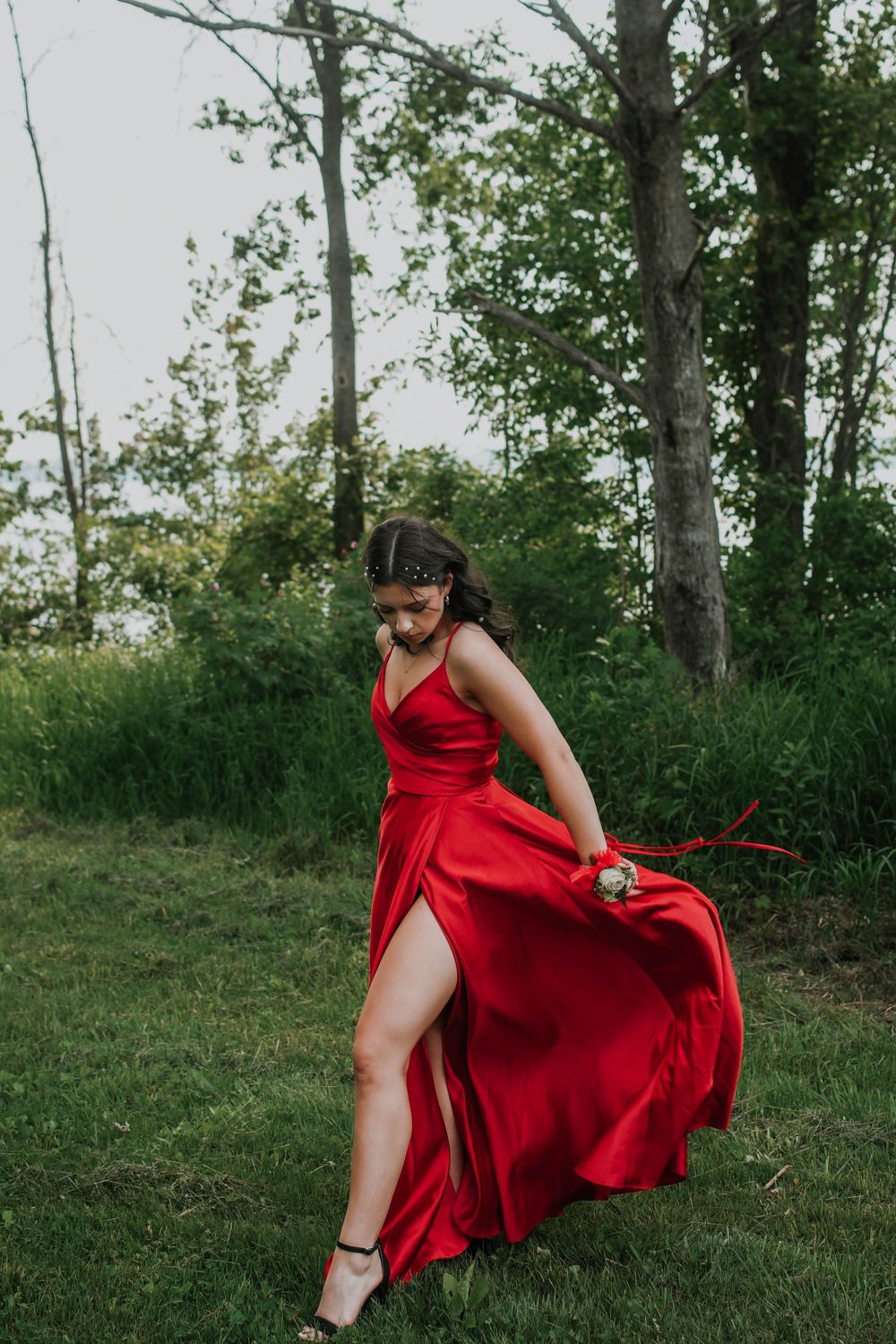 A woman in a flowing red formal dress twirls outdoors in a natural wooded setting on a summer evening.