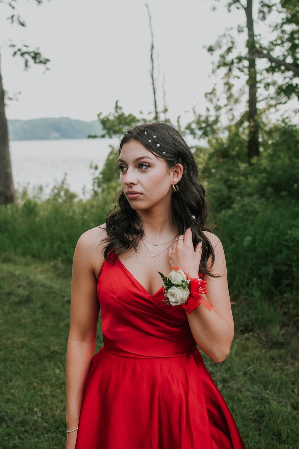 Red prom dress with corsage and hair accessories photographed in natural outdoor setting near a lake.