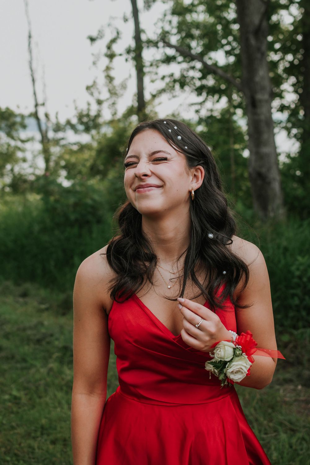 A person in a vibrant red formal dress smiles outdoors with white flowers on their wrist amid a green natural setting.
