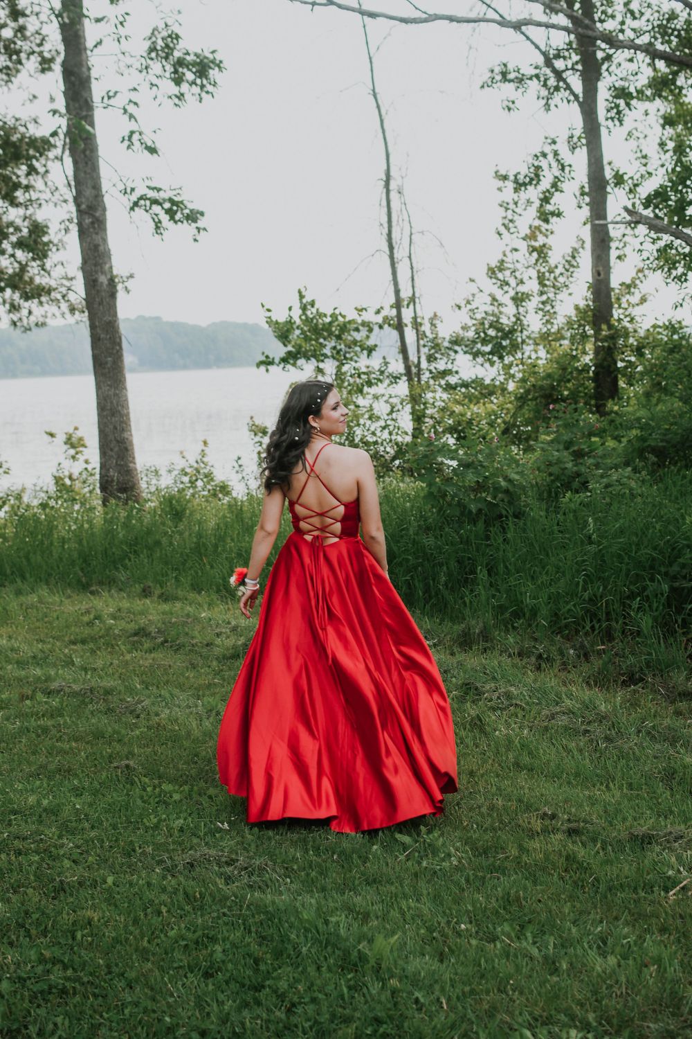 Someone in a flowing red formal gown stands amid trees and greenery near a misty waterfront.