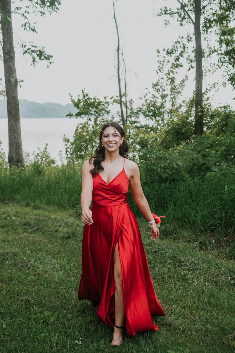 Woman in flowing red gown with thigh-high slit stands on grassy lakeside area surrounded by trees.