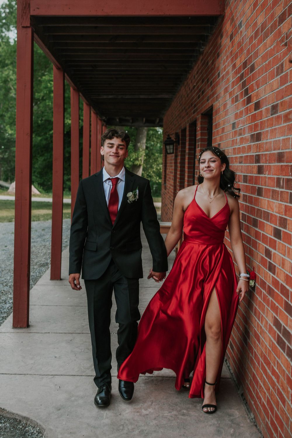 Formal couple walks together along covered brick walkway, wearing black suit and flowing red gown with leg slit.