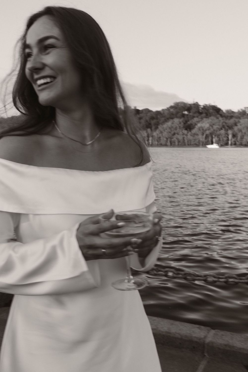 Black and white portrait of woman in off-shoulder dress laughing by the water.