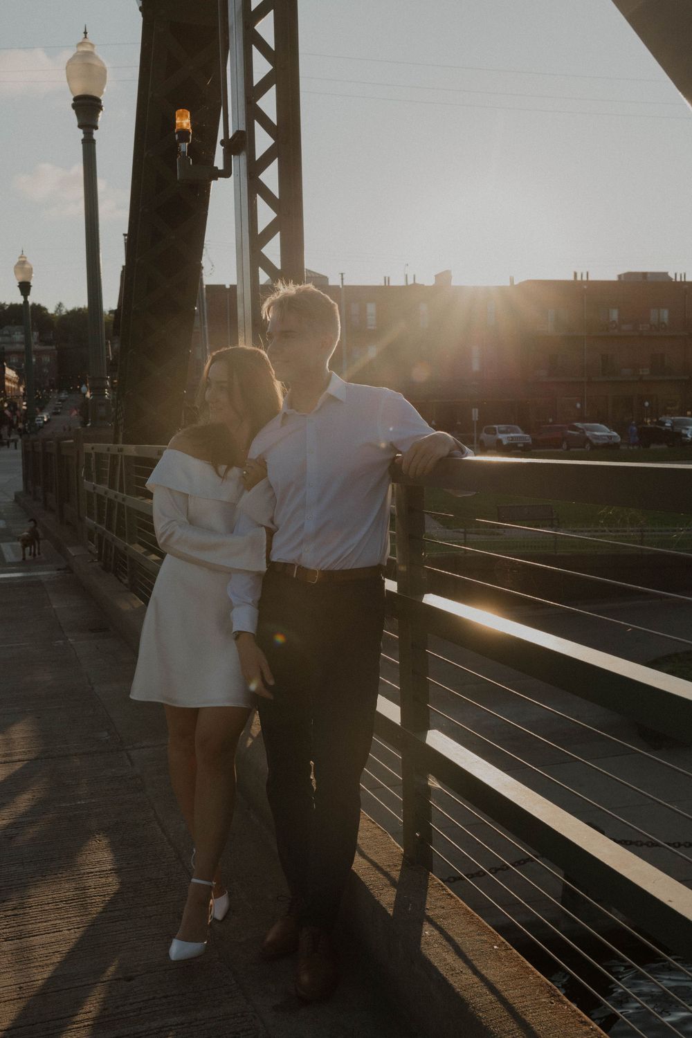 Couple posing on bridge at sunset with urban backdrop and street lamp.